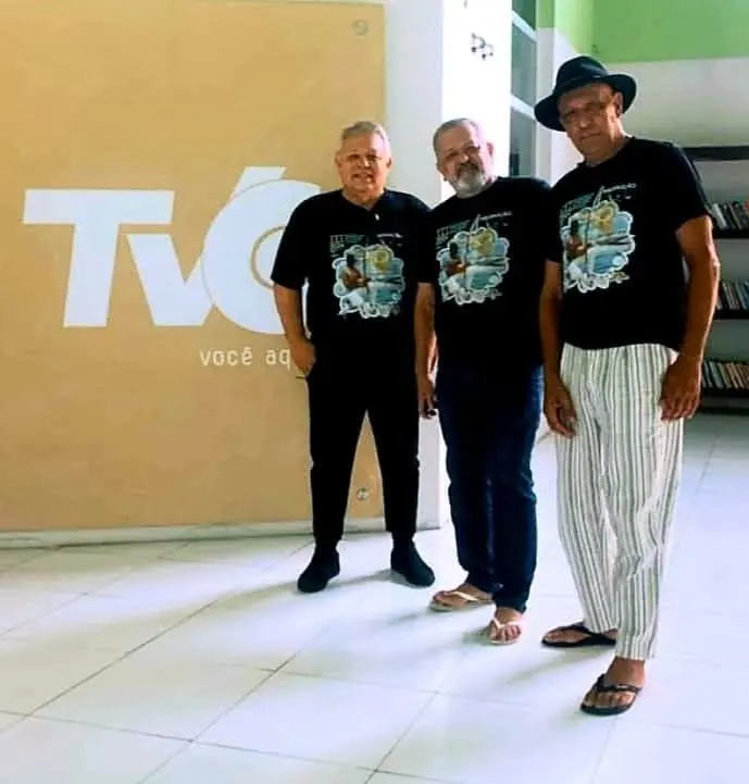 Três mestres de capoeira em frente ao logotipo da TVC